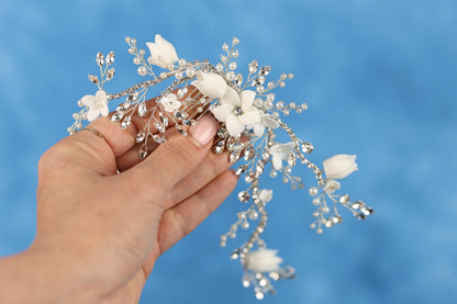 A hand holding a decorative hair accessory with white flowers and silver beads against a blue background.