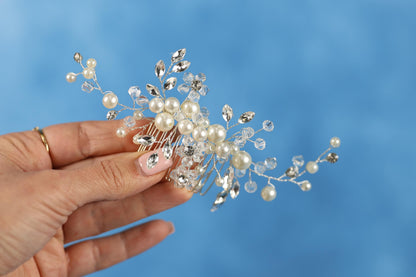 A hand holding a decorative hair accessory with clear gemstones and pearls against a blue background.