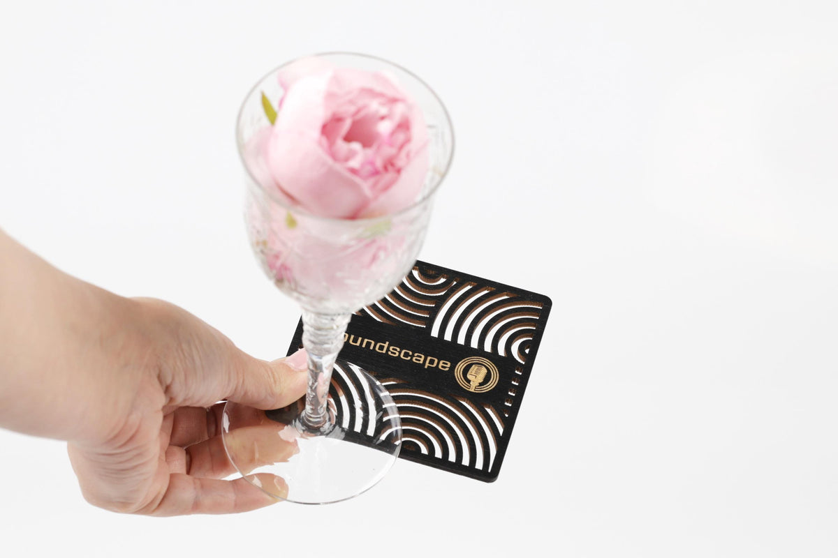A hand holding a glass with a pink flower in it, placed on a coaster with the word "Landscape" printed on it.