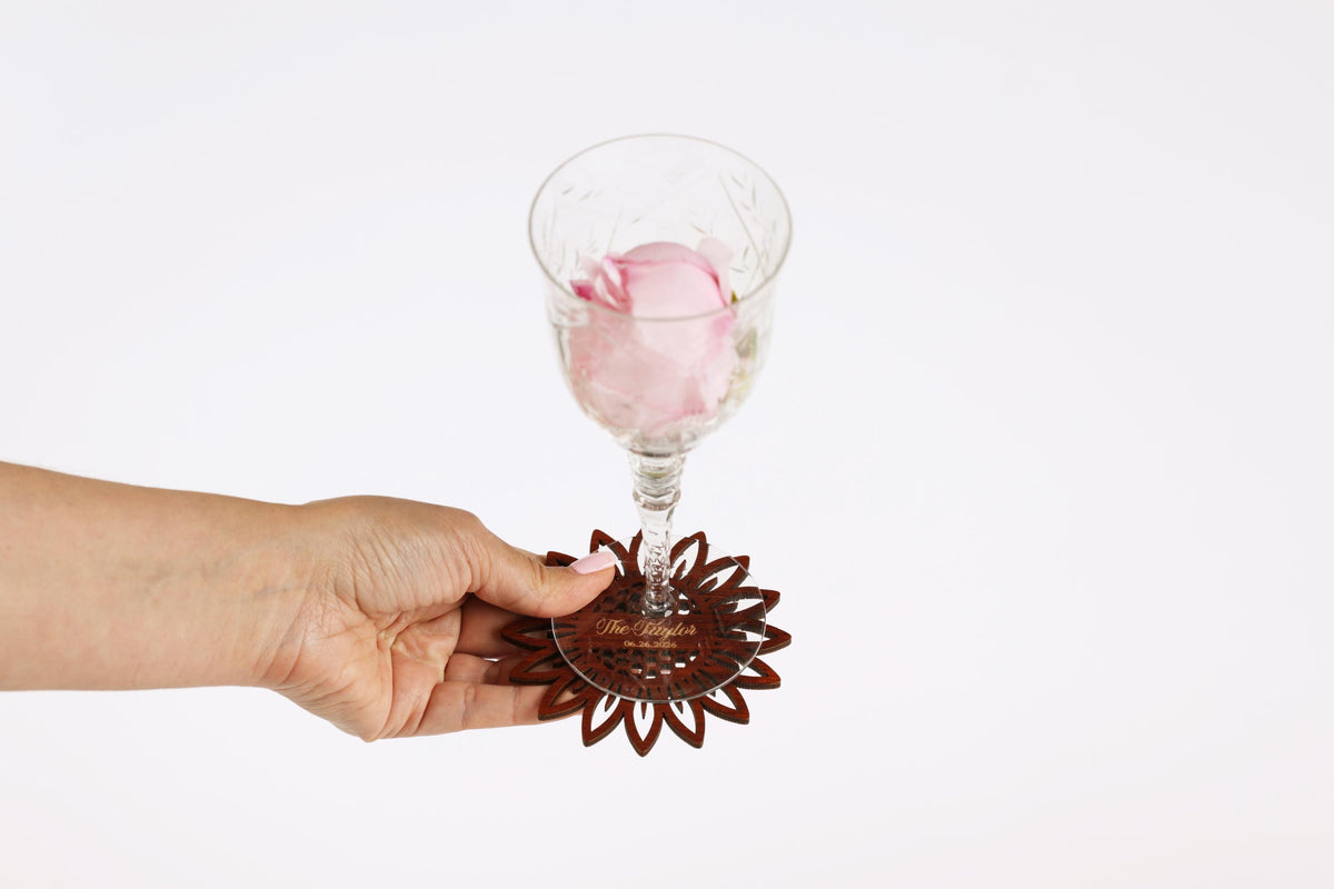 A hand holding a glass with a pink rose inside, and a wooden coaster with a floral design underneath.