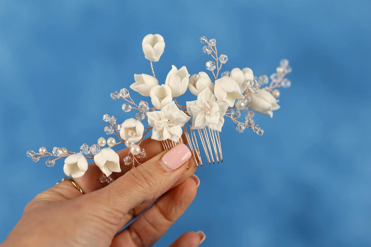 A hand holding a decorative hair accessory with white flowers and crystal embellishments against a blue background.
