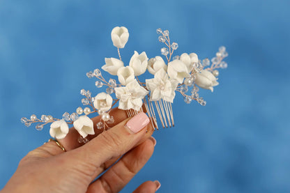 A hand holding a decorative hair accessory with white flowers and crystal embellishments against a blue background.