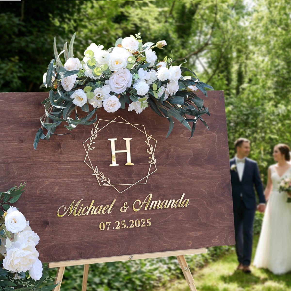 A wooden sign displaying the names "Michael & Amanda" and the date "07.25.2025" is surrounded by a floral arrangement. In the background, a couple is walking together in a garden setting.