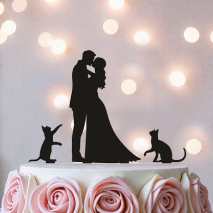A silhouette of a couple embracing, with two cats in the foreground, is displayed on a cake topper against a background of pink roses and white lights.