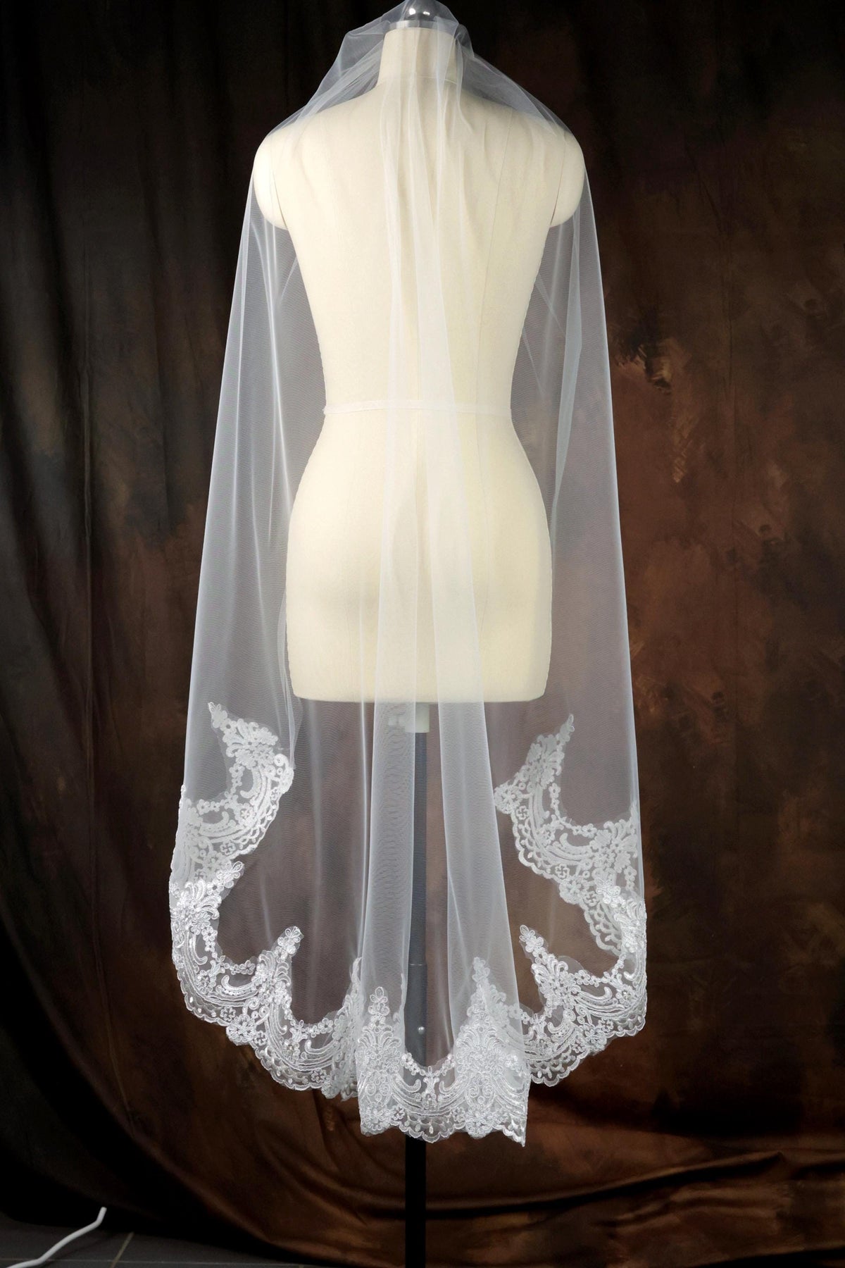 A white wedding veil with a delicate lace trim is displayed on a mannequin against a brown background.
