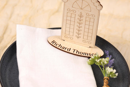 A wooden place card holder with a nameplate sits on a white napkin, accompanied by a small bouquet of flowers.