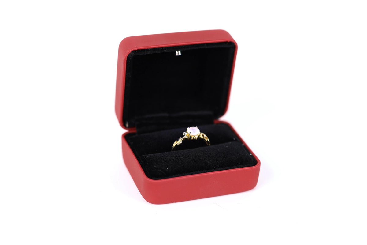A gold ring with a gemstone is displayed in a red jewelry box against a white background.