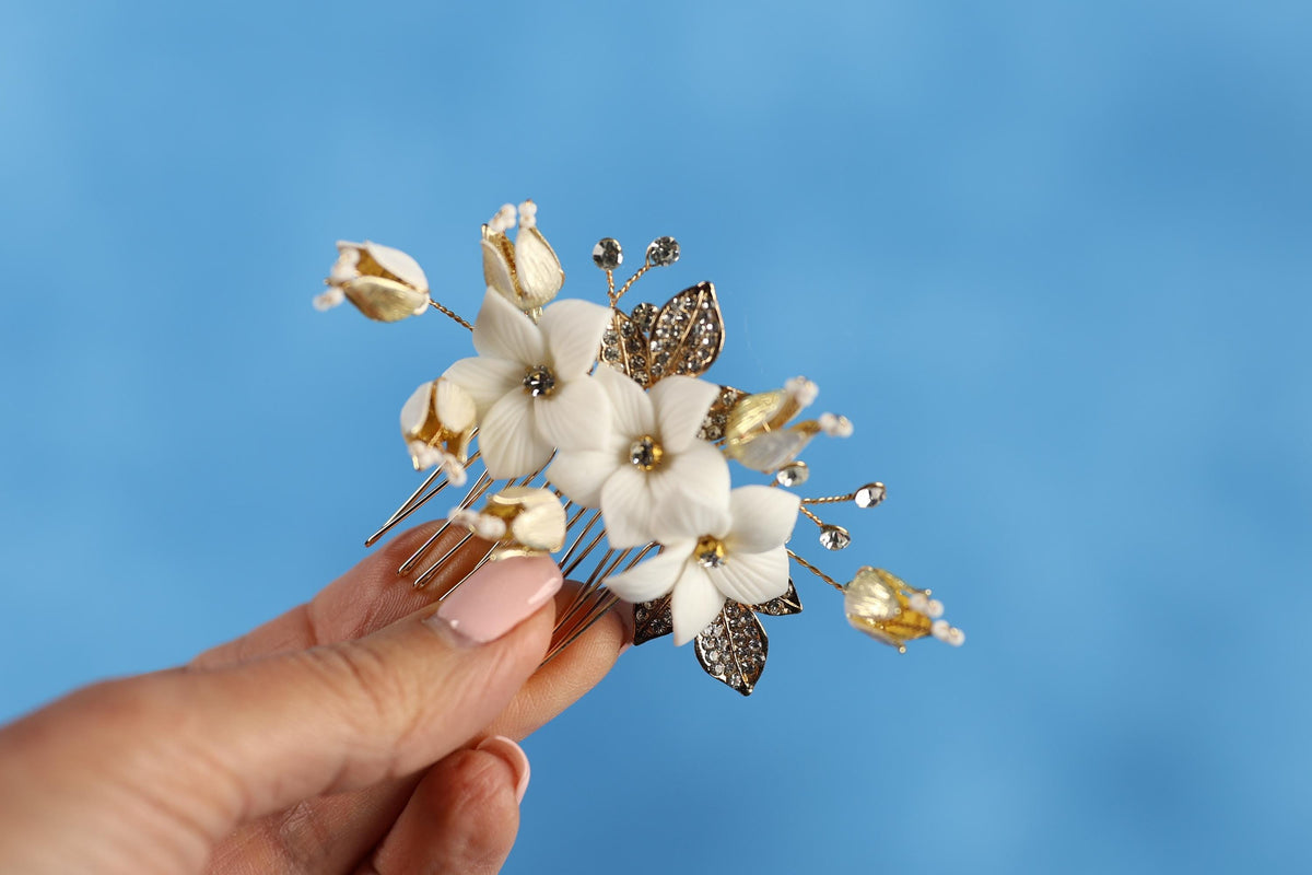 A hand holding a decorative hair clip with white and gold flowers against a blue background.