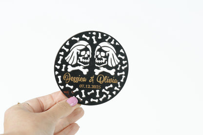 A hand holding a circular object with a black and white design featuring two skull faces and the text " Jessica & Olivia 07.12.2025".