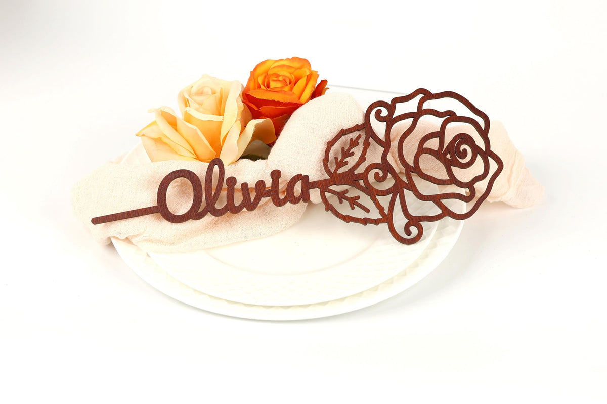 a decorative wooden name tag with the name "Olivia" written on it, placed on a white plate. The name tag is accompanied by two roses, one orange and one yellow, adding a touch of color to the scene.