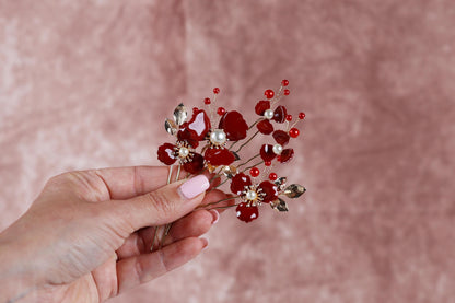 A hand holding a decorative hair clip with red flowers and beads against a pink background.