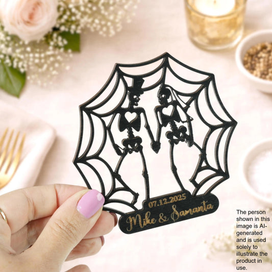 A hand holding a black wedding cake topper featuring a spider web design with two skeletal figures, against a background of flowers and other wedding decorations.