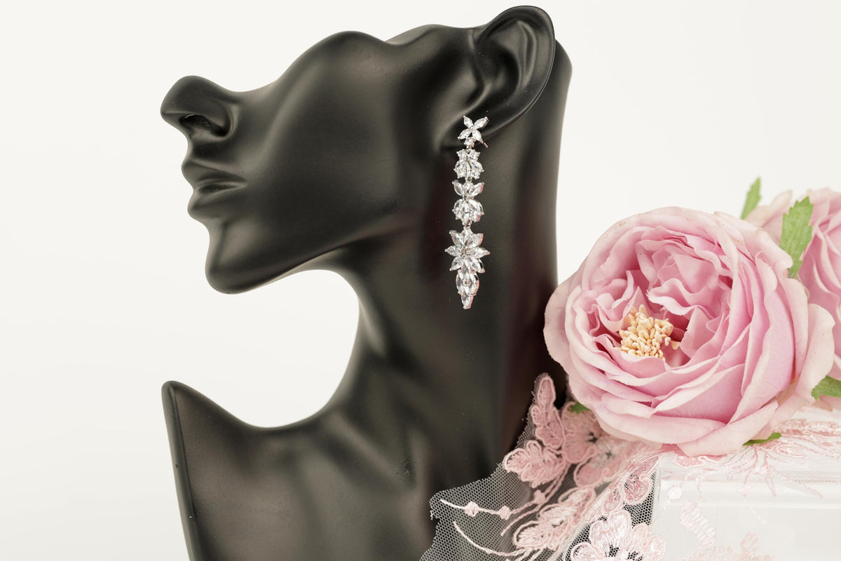 A black mannequin head wearing a pair of diamond earrings with a floral design, accompanied by a pink rose and a delicate lace fabric.