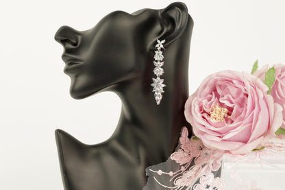 A black mannequin head wearing a pair of diamond earrings with a floral design, accompanied by a pink rose and a delicate lace fabric.