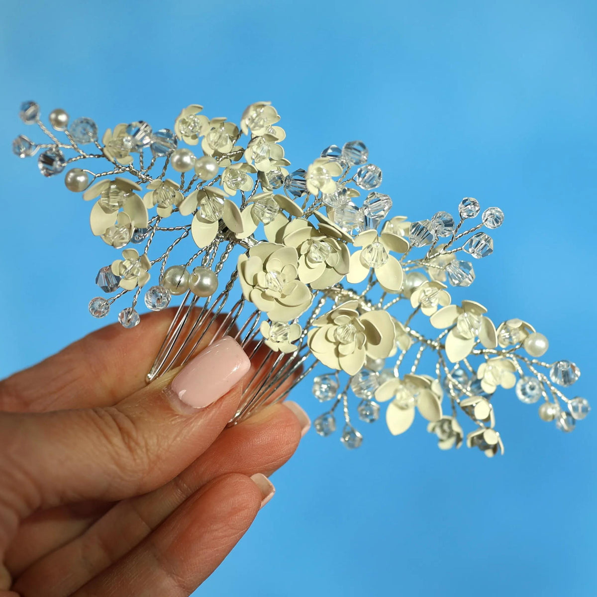 A hand holding a decorative hair accessory with white flowers and clear gemstones against a blue background.