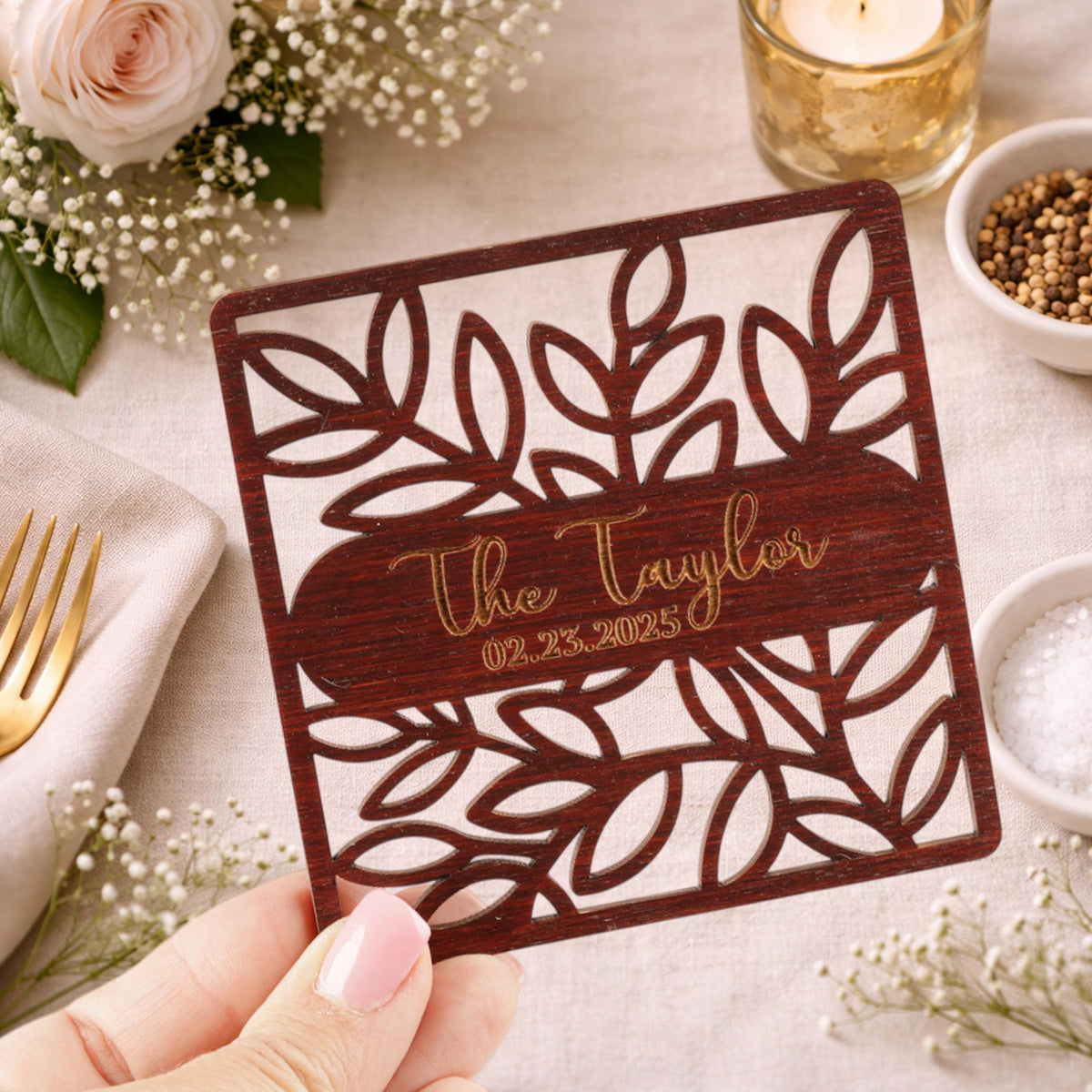 A hand holding a wooden coaster with a floral design and text on it, surrounded by other decorative items such as flowers, a candle, and a bowl of spices.