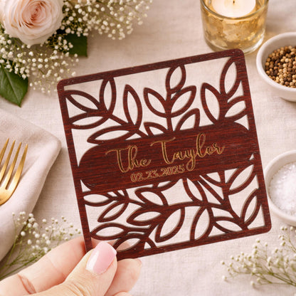 A hand holding a wooden coaster with a floral design and text on it, surrounded by other decorative items such as flowers, a candle, and a bowl of spices.