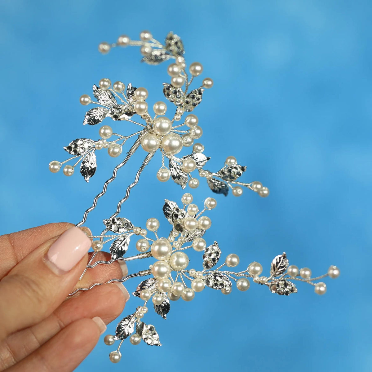 A hand holding a decorative hair clip with small white pearls and silver leaves against a blue background.