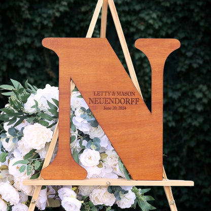 A wooden letter 'N' with the names 'LETTY & MASON NEUENDORFF' engraved on it, standing on a wooden stand with a floral arrangement.