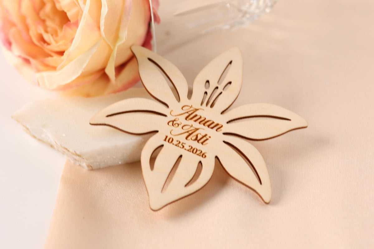 A wooden flower-shaped wedding favor with the names "Aman & Aaliyat" engraved on it, placed on a table next to a pink flower.