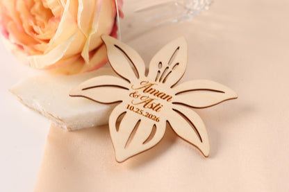 A wooden flower-shaped wedding favor with the names "Aman & Aaliyat" engraved on it, placed on a table next to a pink flower.