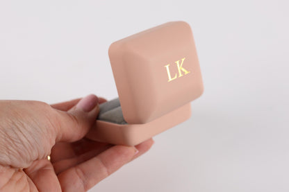 A hand holding a pink jewelry box with a gold "LK" monogrammed on it.