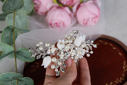A hand holding a decorative hair accessory with white flowers and pearls, surrounded by pink roses and greenery.