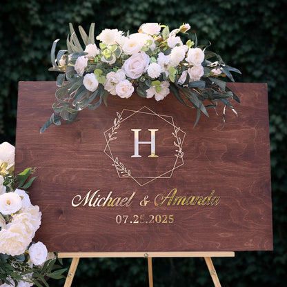 A wooden sign with a floral arrangement and a large letter "H" on it, displaying the names "Michael & Amanda" and the date "07.25.2025".