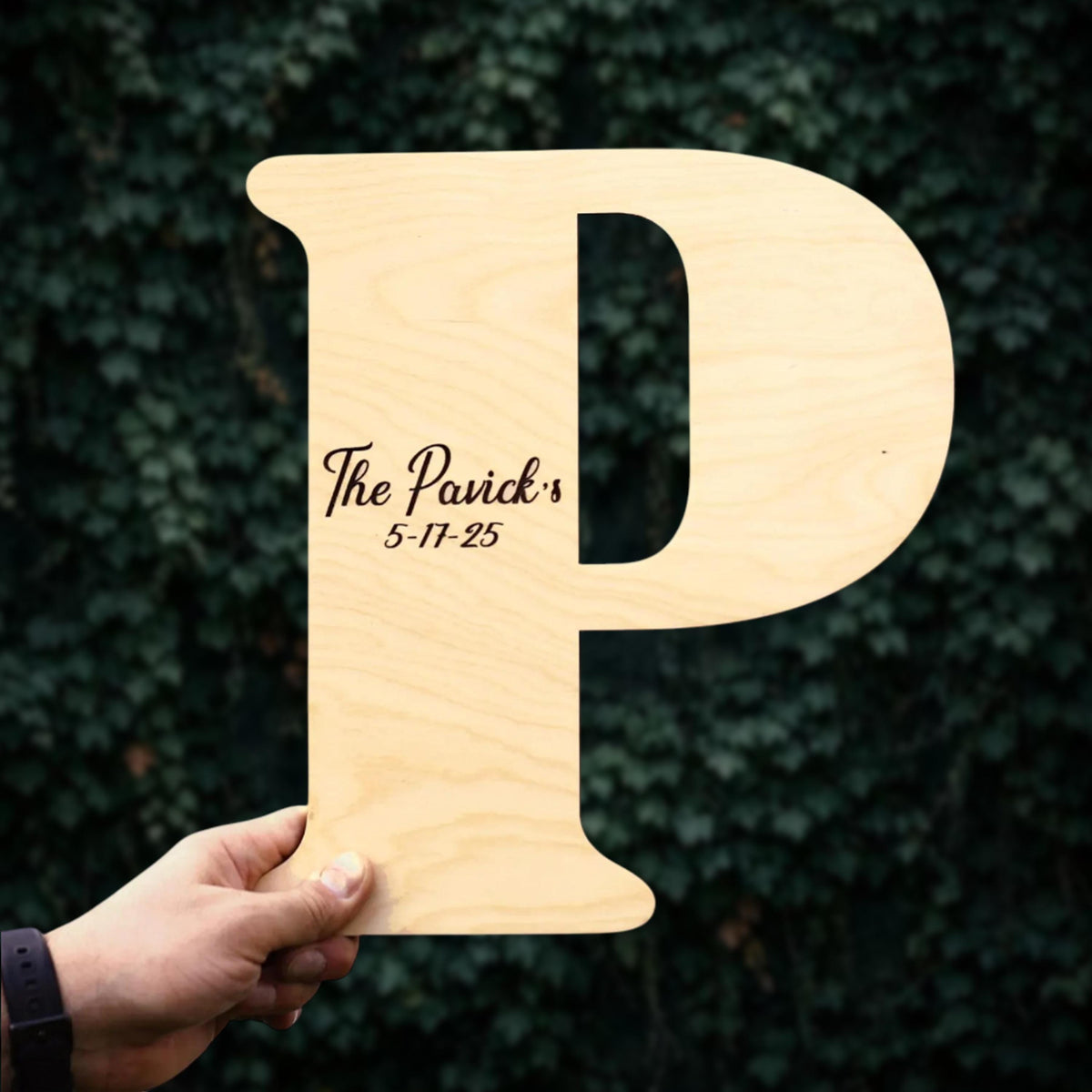 A person's hand holding a wooden letter 'P' with text on it against a background of green ivy leaves.