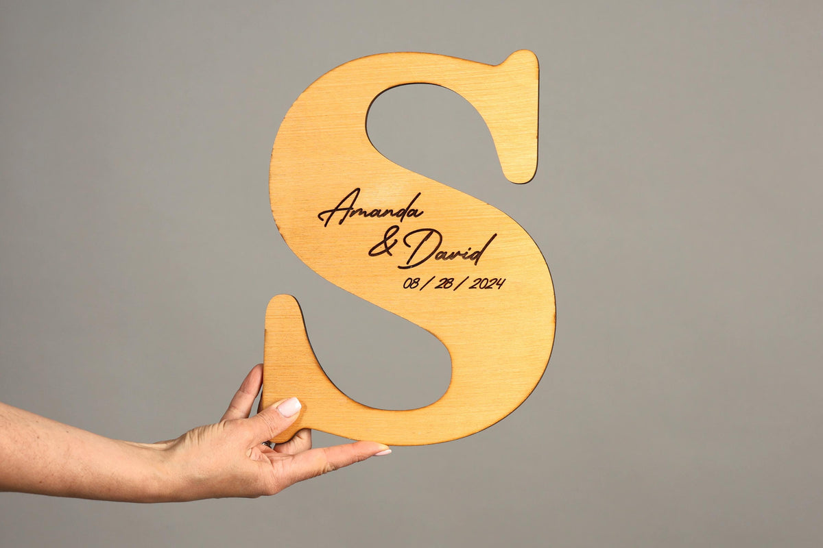 A hand holding a wooden letter 'S' with the names 'Amanda & David' and the date '08/28/2024' engraved on it.