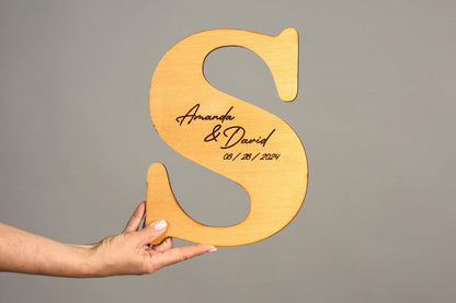 A hand holding a wooden letter 'S' with the names 'Amanda & David' and the date '08/28/2024' engraved on it.