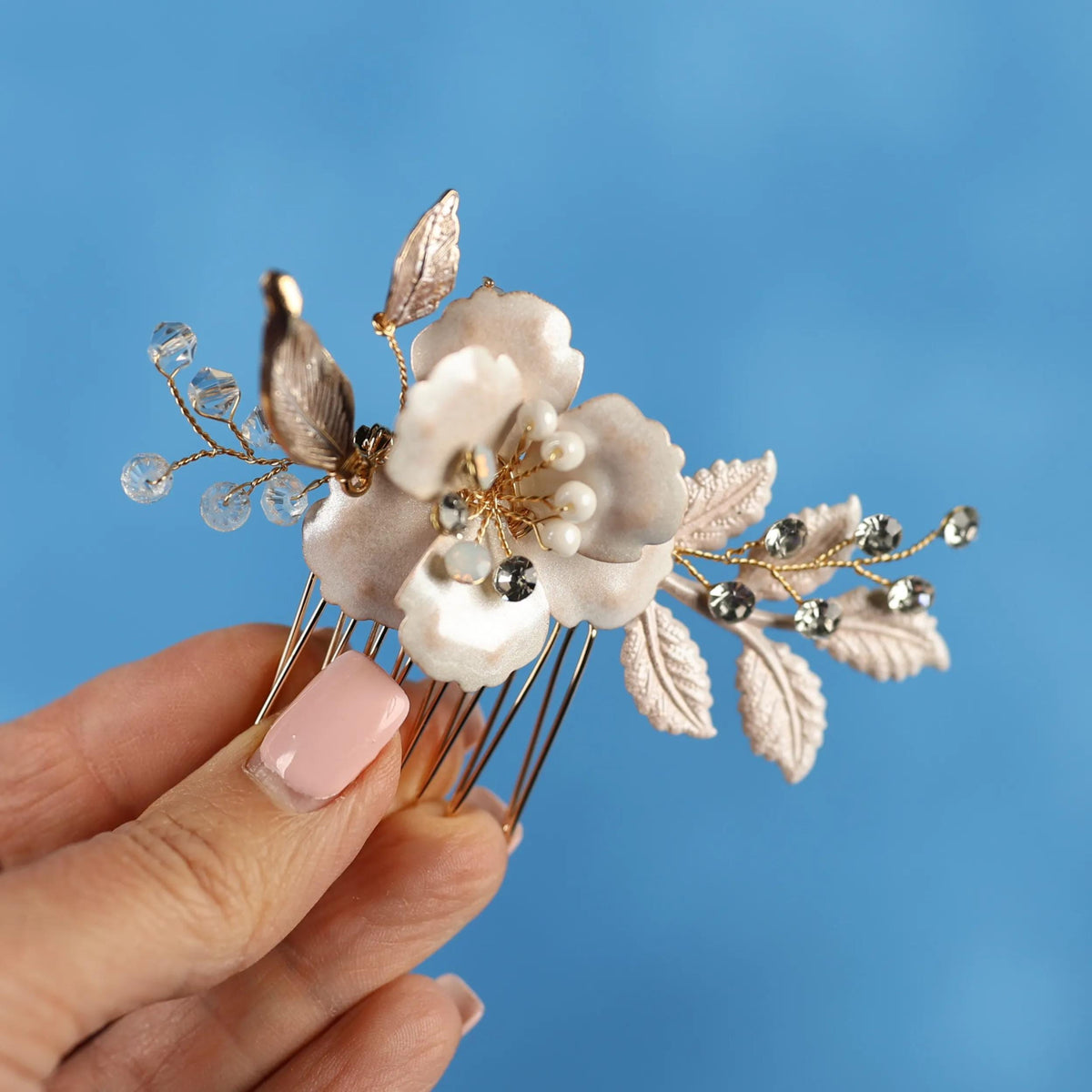 A hand holding a decorative hair clip with a white flower and leaves against a blue background.