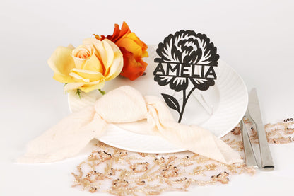 a white plate with a floral design, a ribbon, and a nameplate with a flower. There are also some flowers and a beaded necklace on the table.