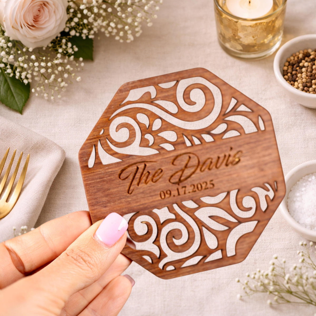 A hand holding a wooden coaster with a floral design, surrounded by other decorative items such as a rose, a candle, and a bowl of spices.