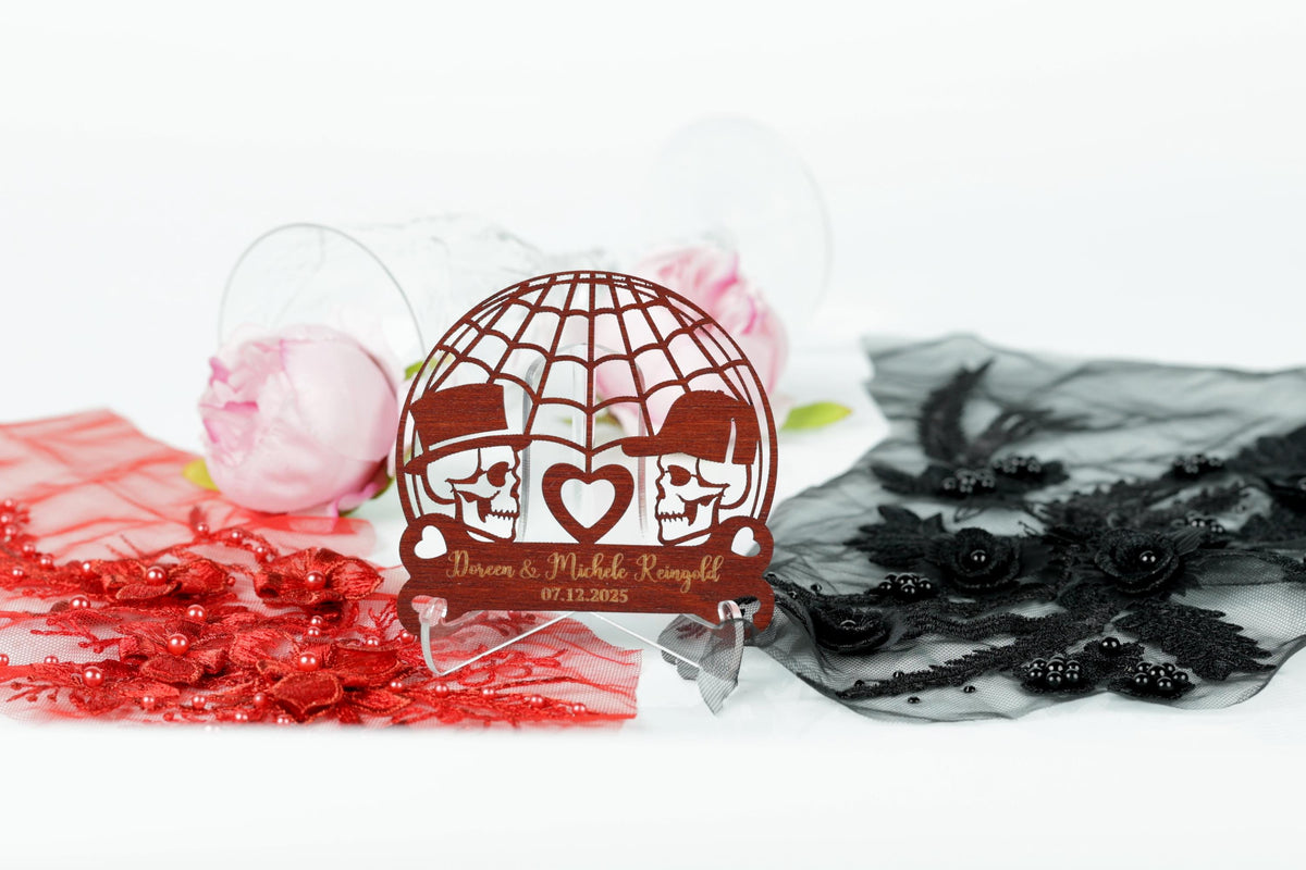 A wooden wedding card with a design featuring two skull faces and a heart, placed on a stand surrounded by various decorative elements such as roses, lace, and a red fabric.