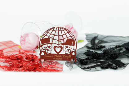 A wooden wedding card with a design featuring two skull faces and a heart, placed on a stand surrounded by various decorative elements such as roses, lace, and a red fabric.
