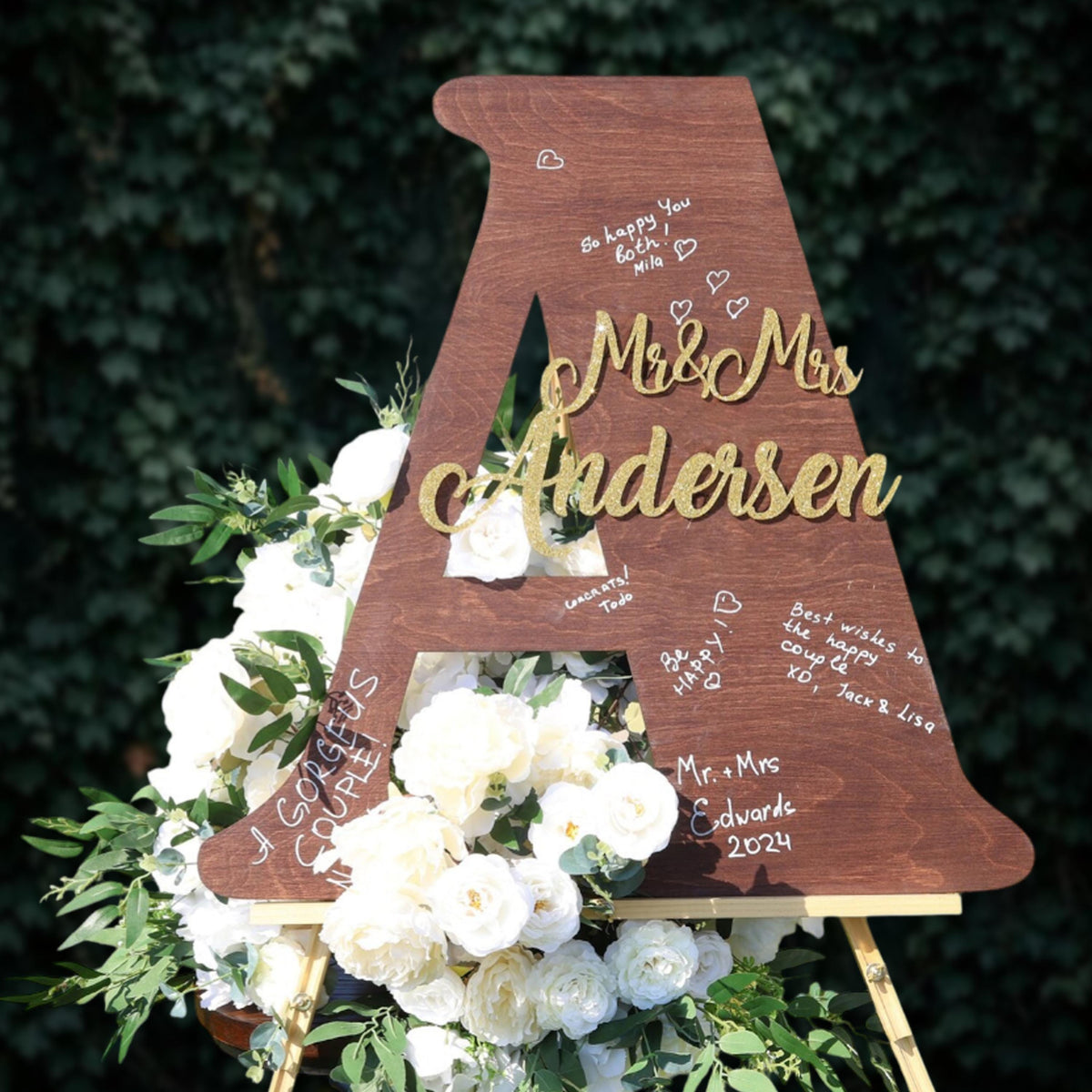 A wooden letter A with the names "Mr & Mrs Andersen" written on it, surrounded by white flowers and greenery.