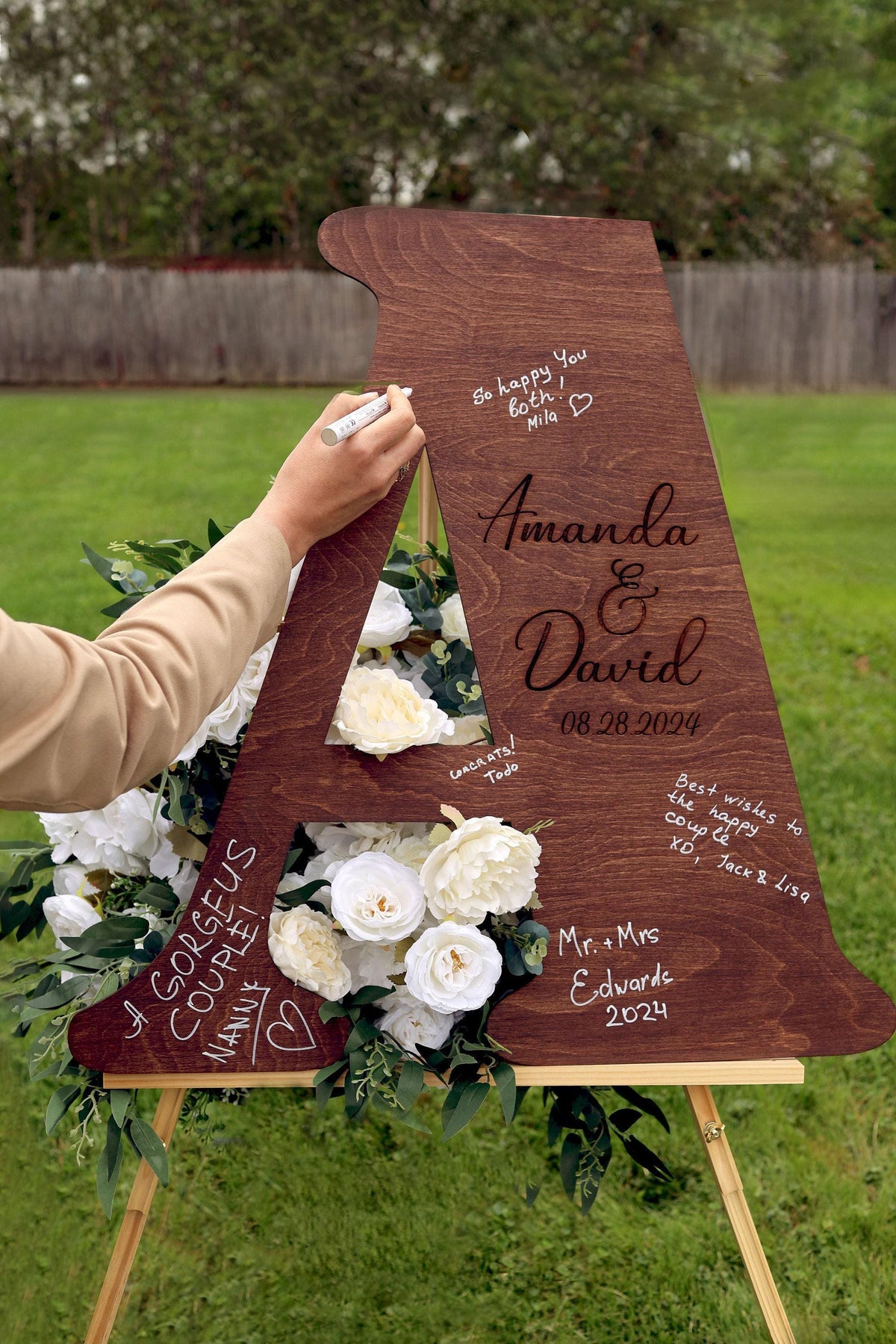 A person is writing on a wooden sign that says "Amanda & David" in a grassy outdoor setting.