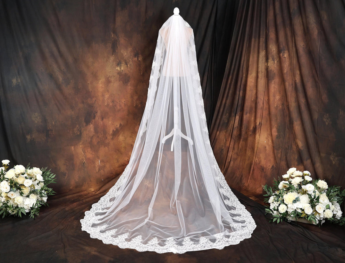 A white wedding veil with a lace trim is displayed in front of a brown curtain, accompanied by a bouquet of white flowers on either side.