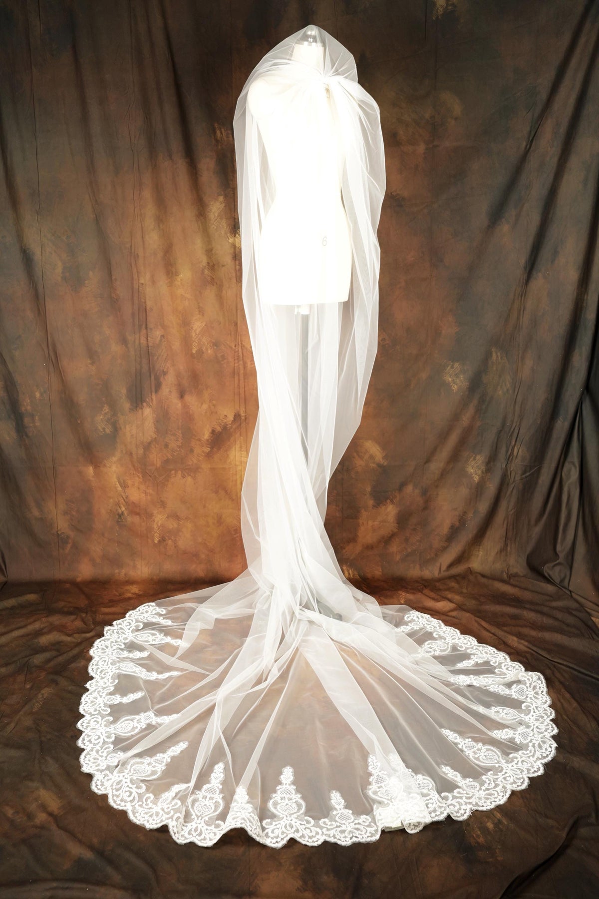 A white wedding veil with a long, flowing train and a lace trim is displayed on a mannequin against a brown background.