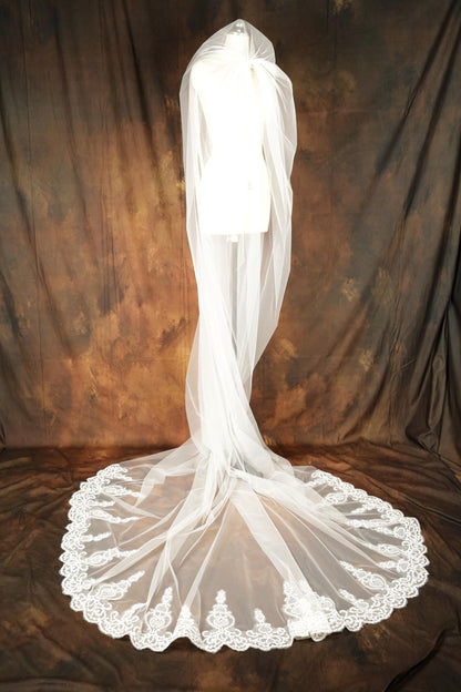 A white wedding veil with a long, flowing train and a lace trim is displayed on a mannequin against a brown background.