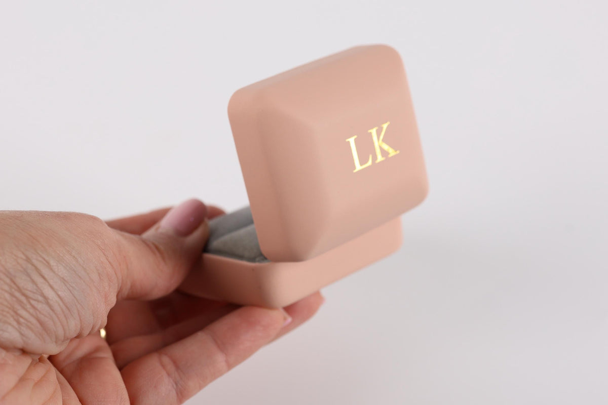 A hand holding a pink jewelry box with a gold "LK" monogrammed on it.