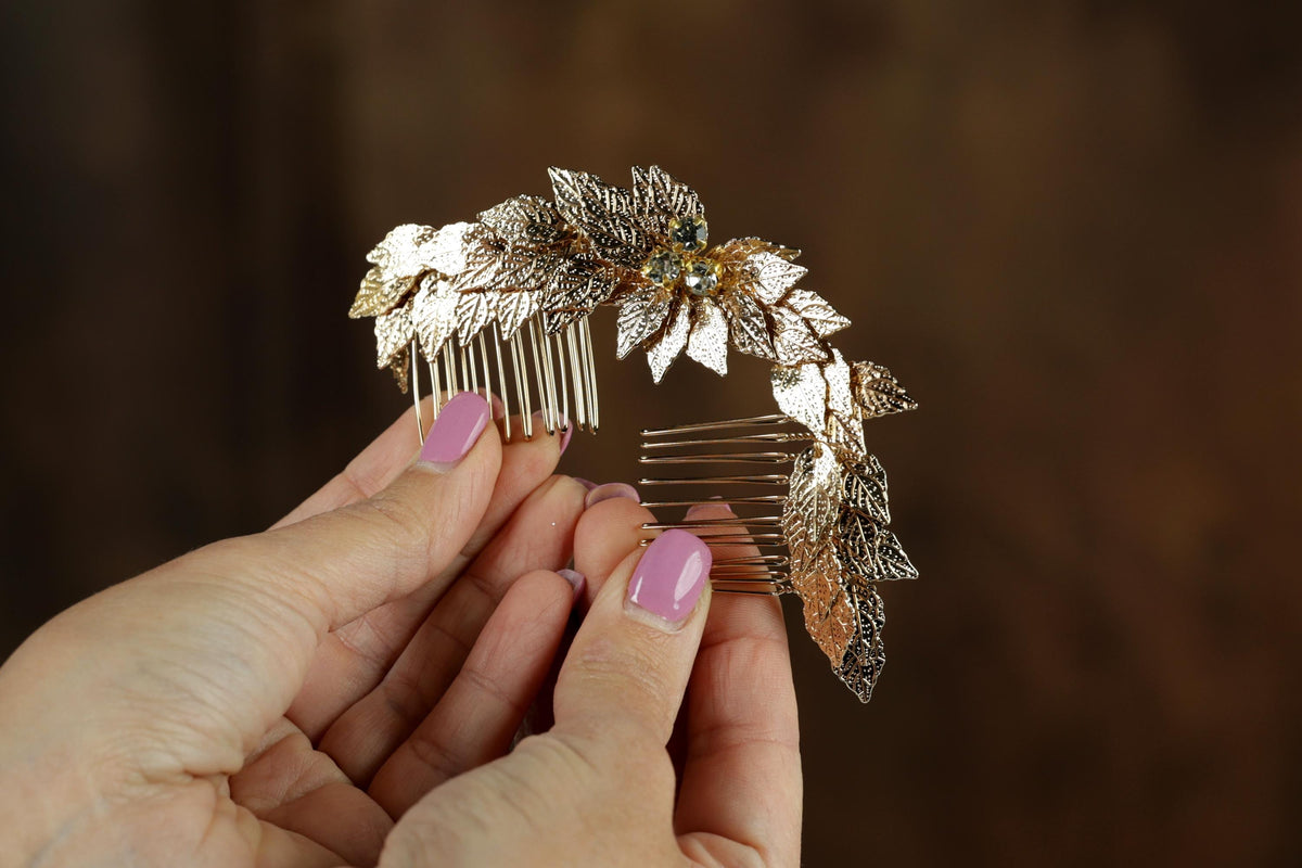 A close-up image of a person's hands holding a decorative gold hair accessory with leaves and gemstones.