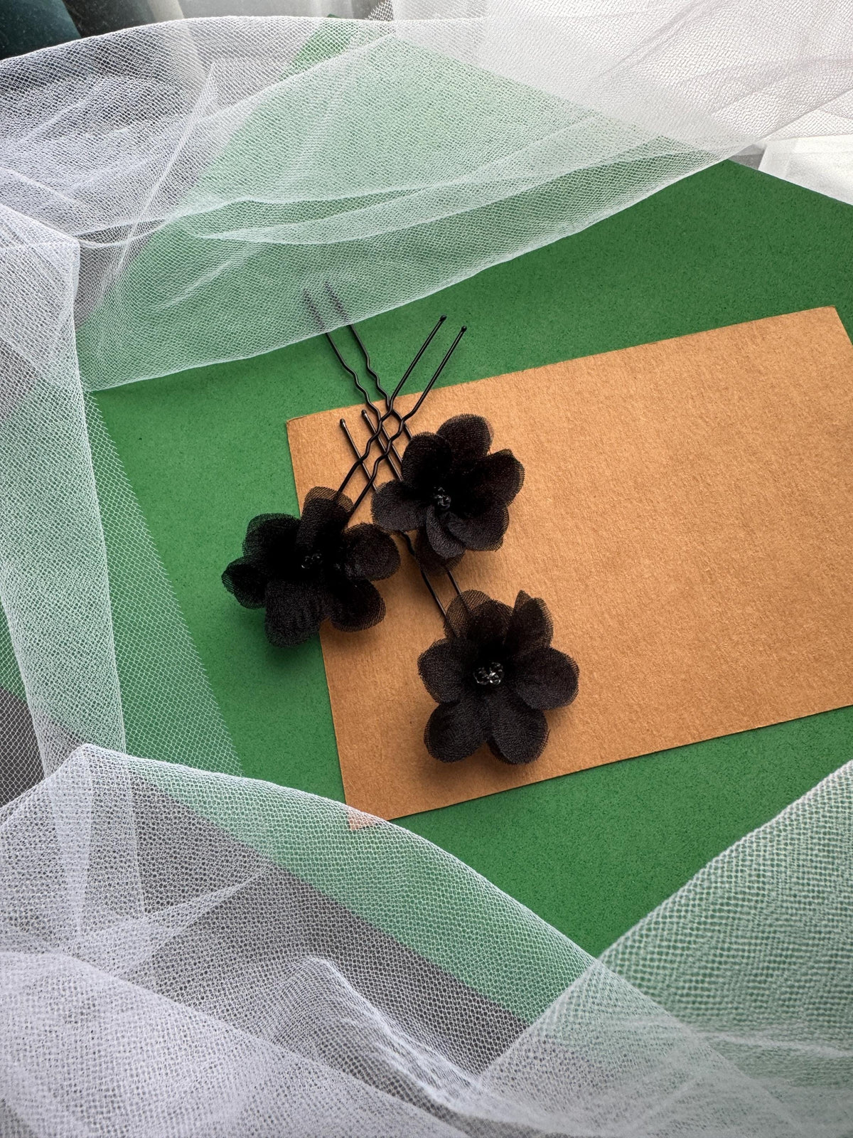 three black flower-shaped hair clips arranged on a green background, with a white net or veil draped over the scene.