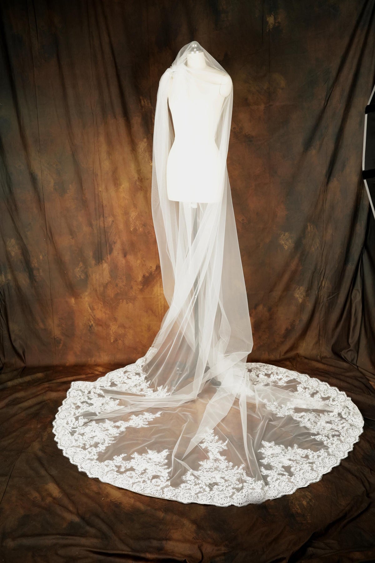 A white wedding dress with a long, flowing veil is displayed on a mannequin against a brown background.