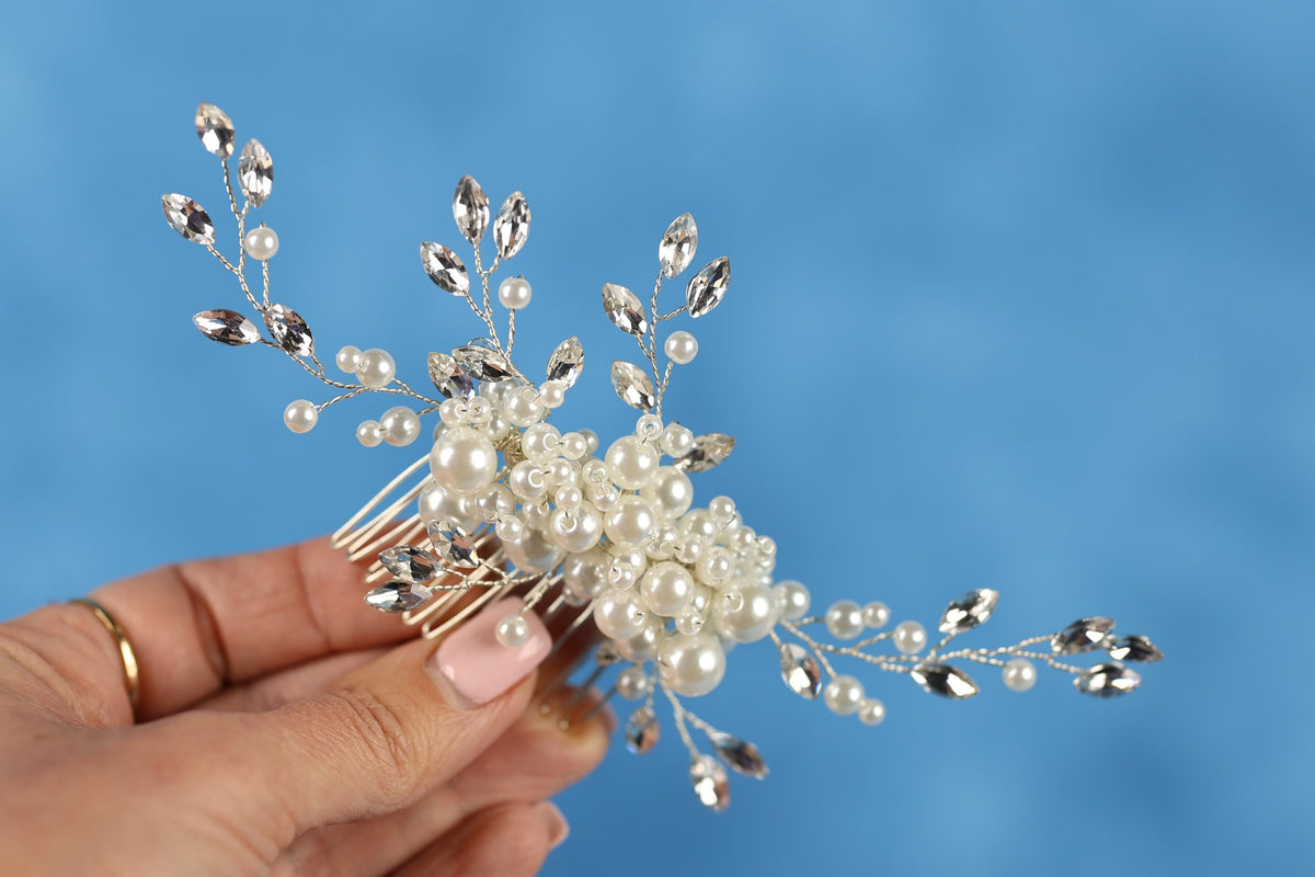 A hand holding a decorative hair clip with white pearls and silver beads against a blue background.