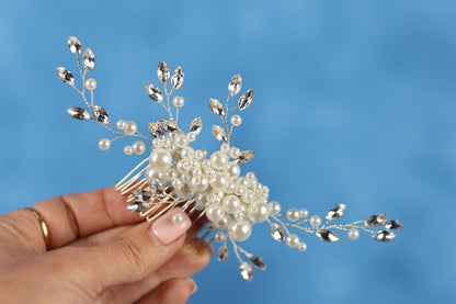 A hand holding a decorative hair clip with white pearls and silver beads against a blue background.