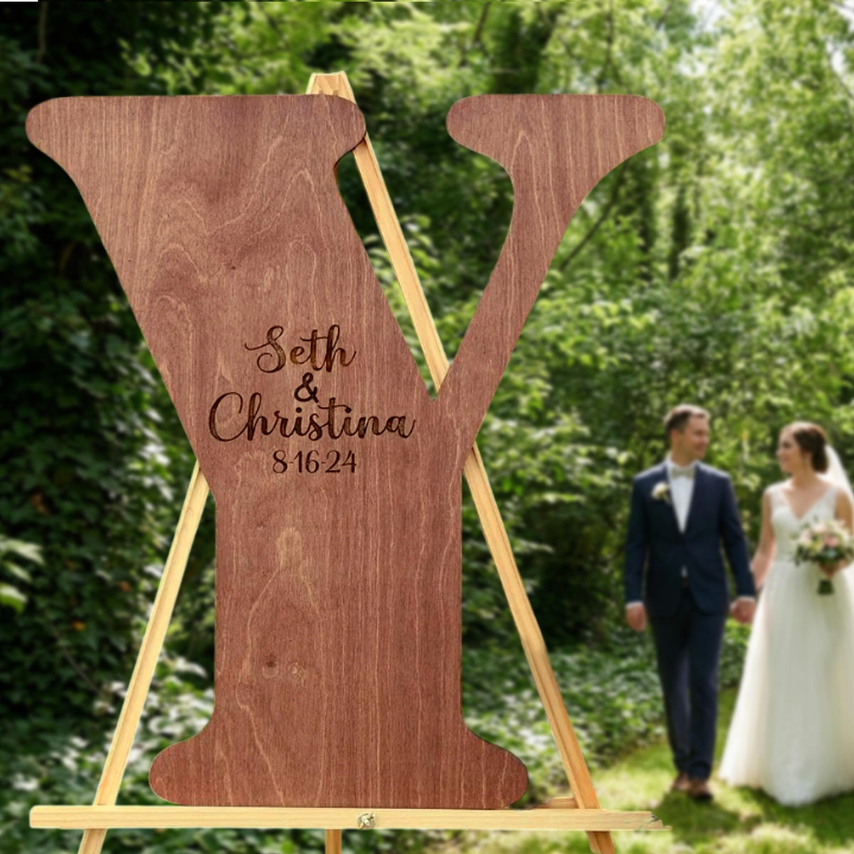 A wooden sign with the names Seth and Christina, and the date "81624", stands in a lush green forest. A couple is walking in the background, suggesting a wedding celebration.