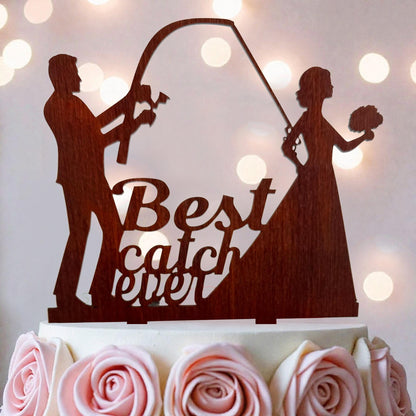 A wooden cake topper with silhouettes of a man and a woman, along with the text "Best catch ever" written on it.