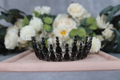 A black ornate crown with black gemstones is displayed in the foreground, with a bouquet of white flowers and greenery in the background.